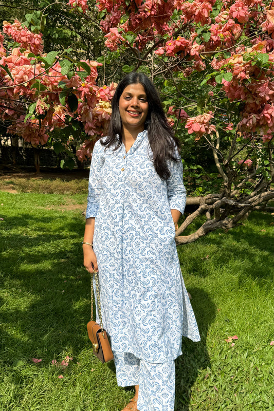 A woman standing in a garden wearing a white printed pure cotton kurta set with floral patterns, paired with matching pants, in a sunny setting with pink flowers in the background.