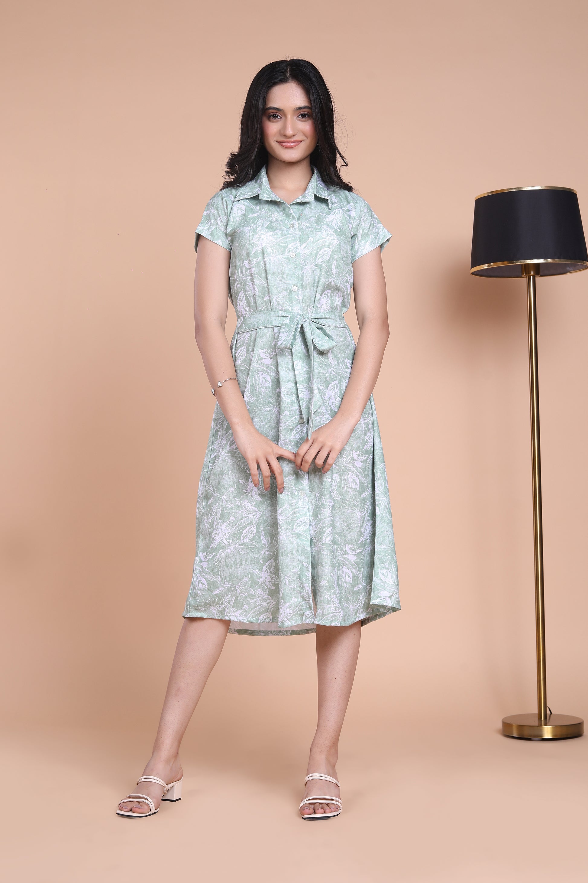 A woman standing next to a lamp post, wearing a green floral midi dress with a belt, in a indoor setting.
