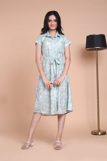 A woman standing next to a lamp post, wearing a green floral midi dress with a belt, in a indoor setting.
