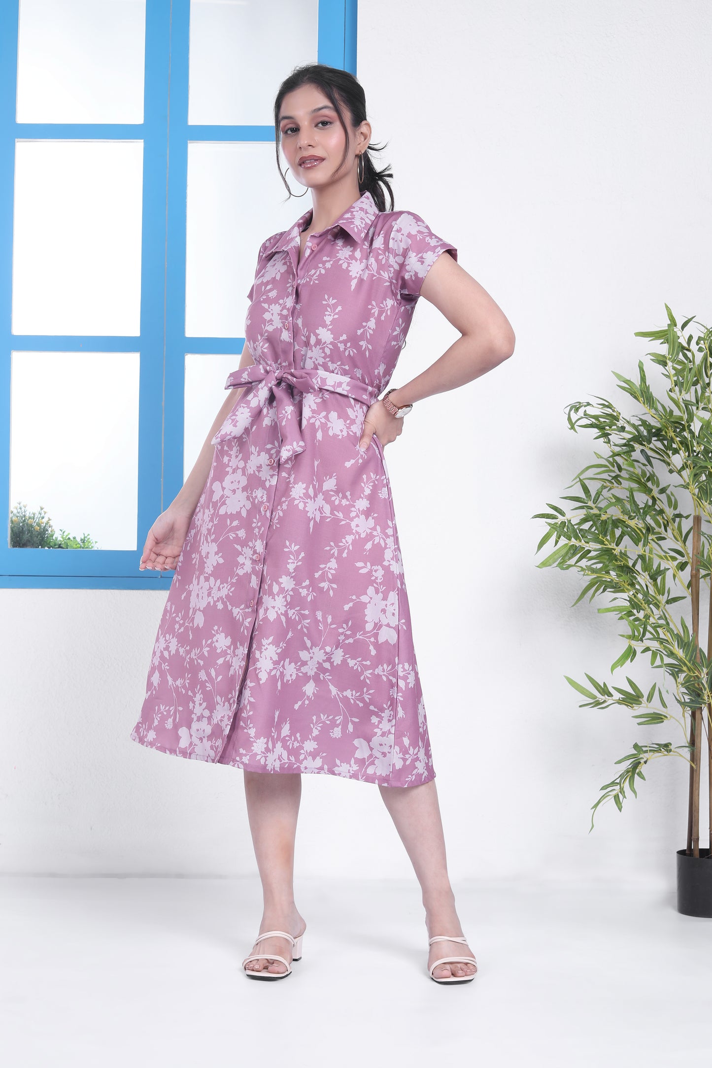 A woman standing in a room with a blue door and window, wearing a pink floral midi dress with a belt, paired with open-toe heeled sandals.