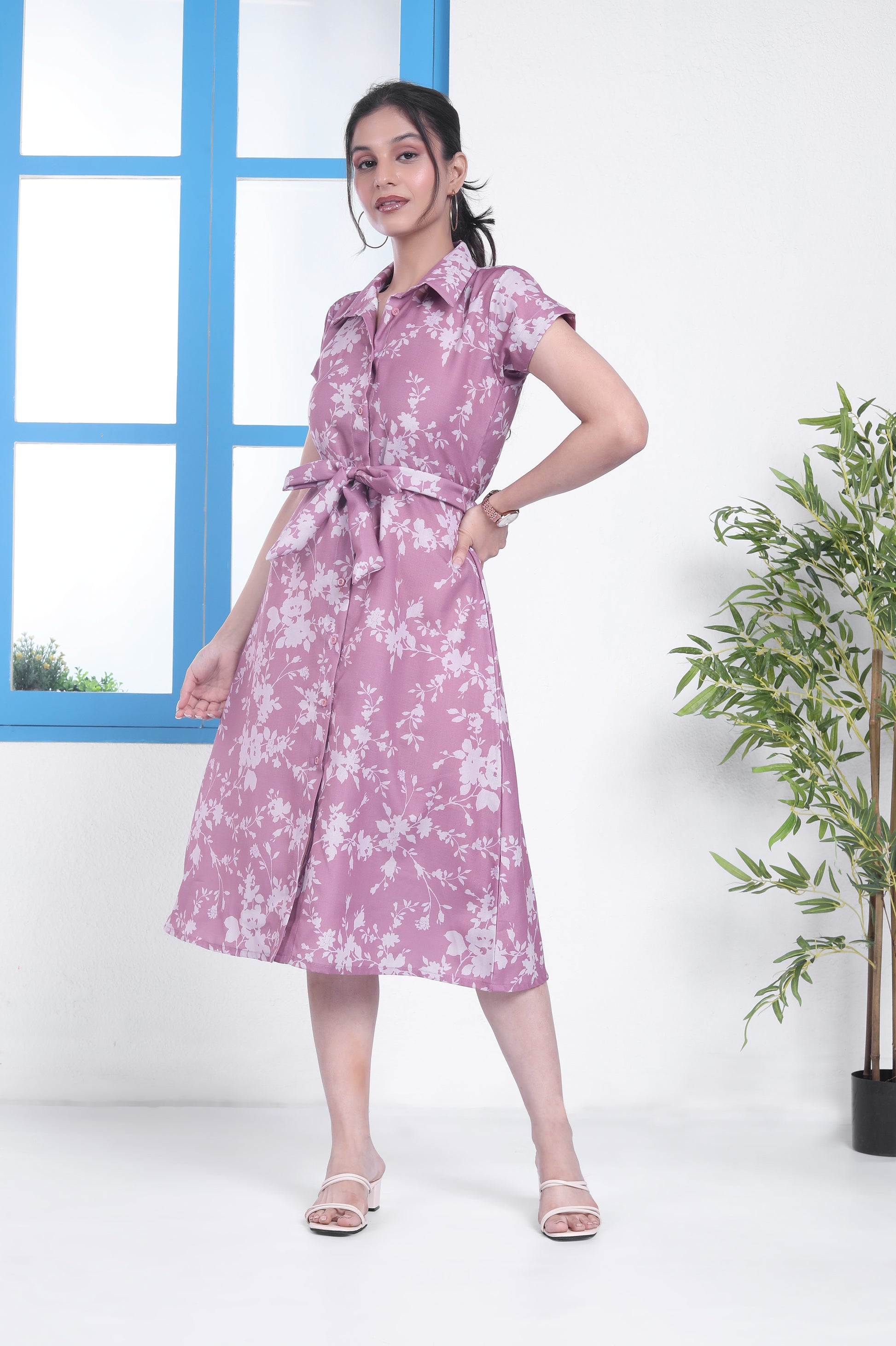 A woman standing in a room with a blue door and window, wearing a pink floral midi dress with a belt, paired with open-toe heeled sandals.